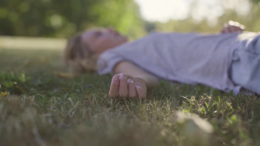 A young girl with blonde hair lays on her back atop grass while in the shade of nearby trees