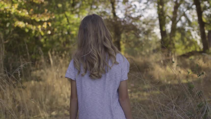A young girl walks slowly along an outdoor trail lined with trees and dried grass