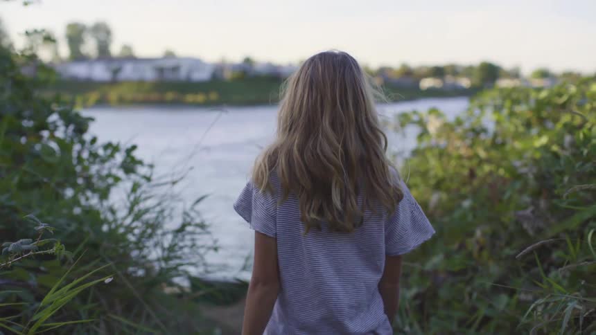 A young girl with blonde hair slowly walks along dirt trail leading to a riverbank