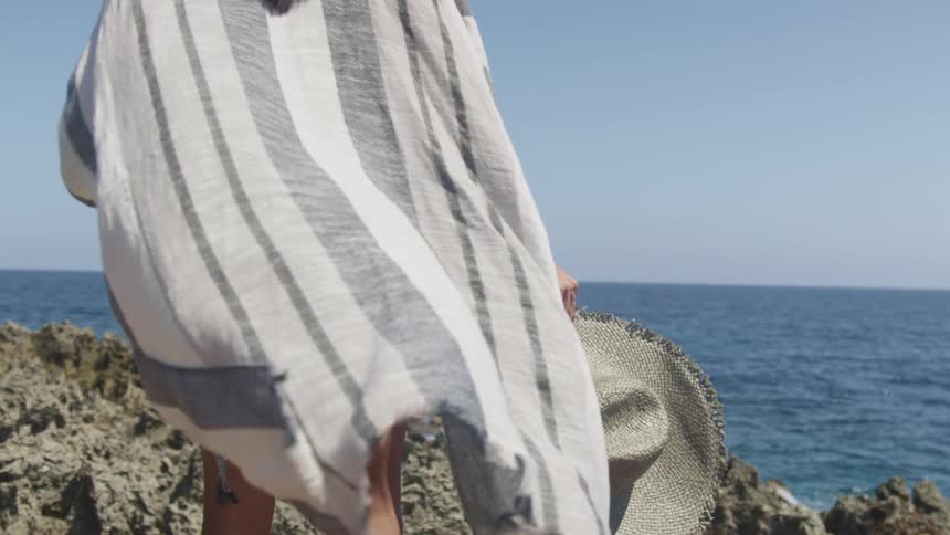 A woman holding her sun hat stands outdoors while looking out upon the ocean