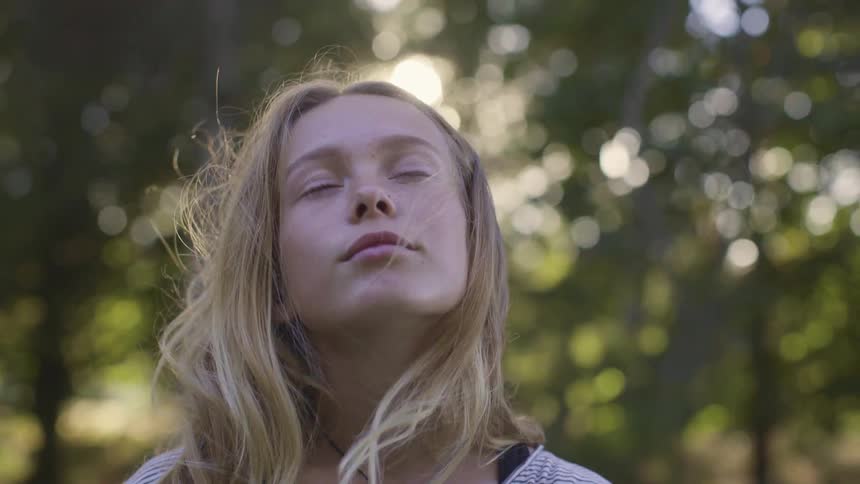 A girl stands outside with her eyes closed surrounded by trees