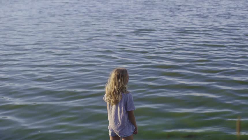 A blonde girl stands at the edge of a lake looking out during the day