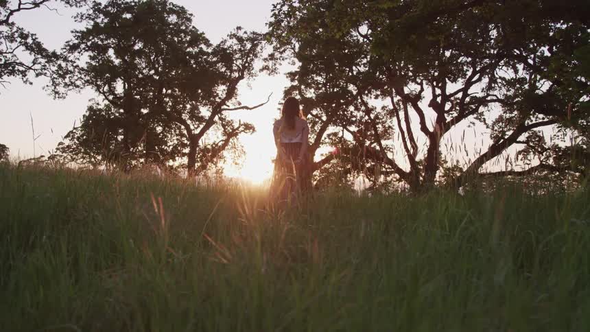 Girl with long hair wearing long skirt standing with her hands behind her back as she watches sun set between the trees