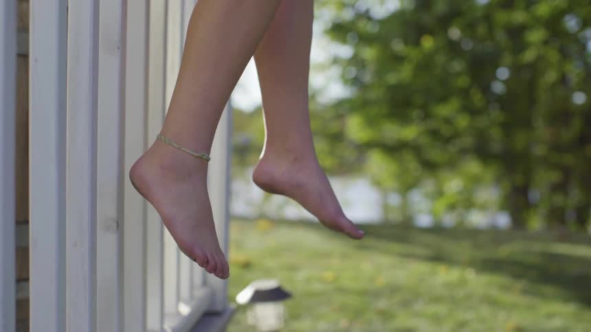 A girl sits on a white fence with bare legs and feet dangling outdoors with trees and grass during the day