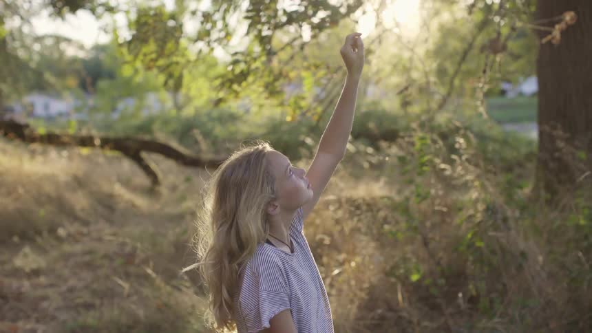 Girl with long blonde hair has her arm in the air reaching for something on a tree branch in the woods