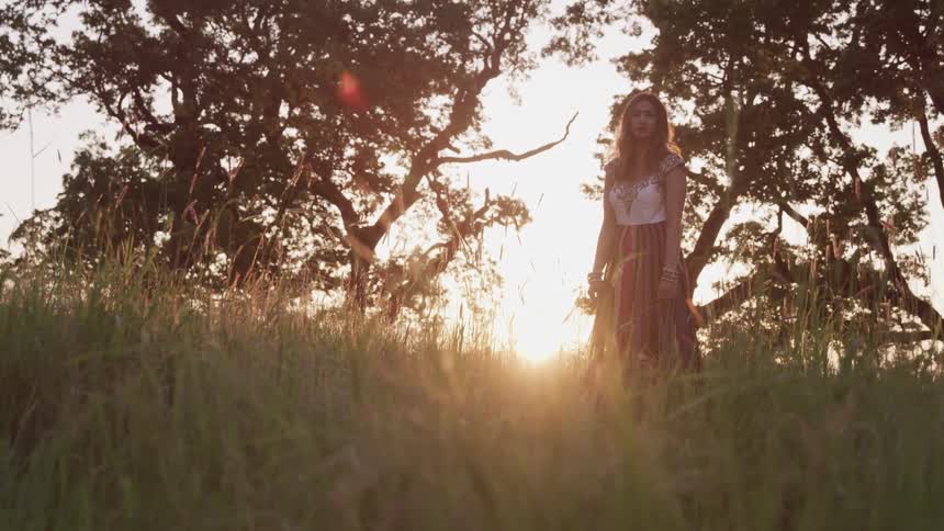 Young woman wearing a dress stands on the grass of a field at sunset