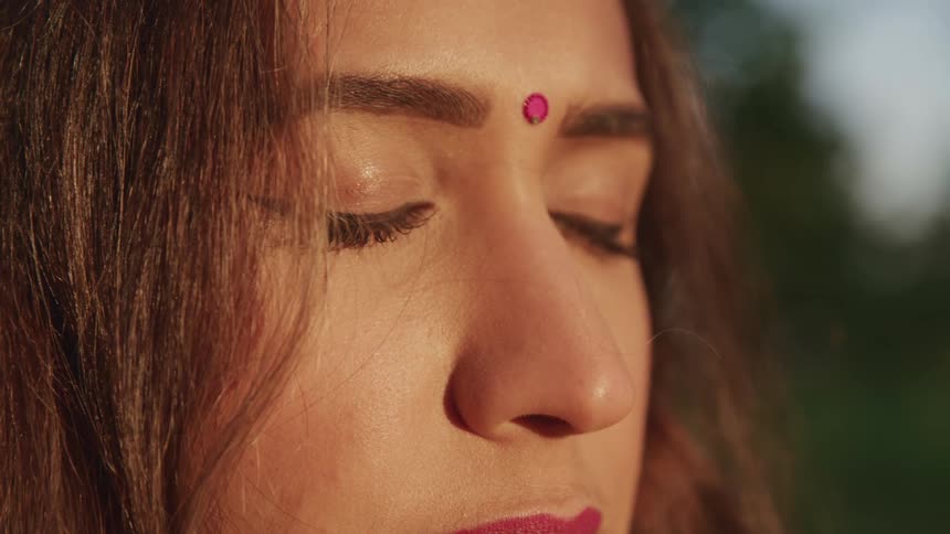 Woman with a pink bindi on her head closing her eyes in a meditative state outdoors