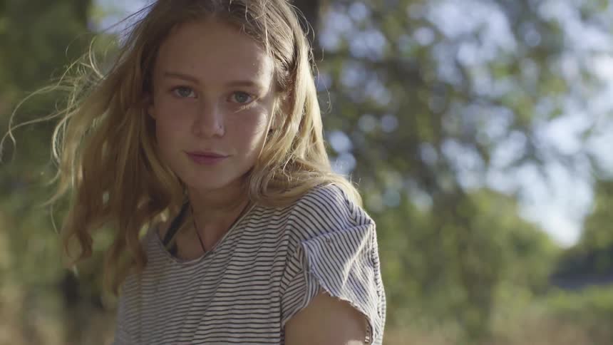 A girl sitting outdoors on a sunny day raising her gaze to look into the camera as wind blows through her hair