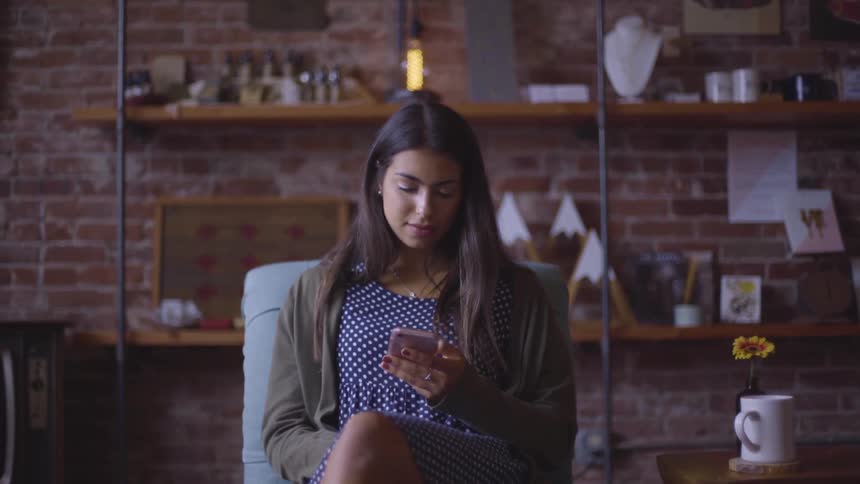 A woman sits in a chair in her apartment in front of a brick wall and uses a smartphone