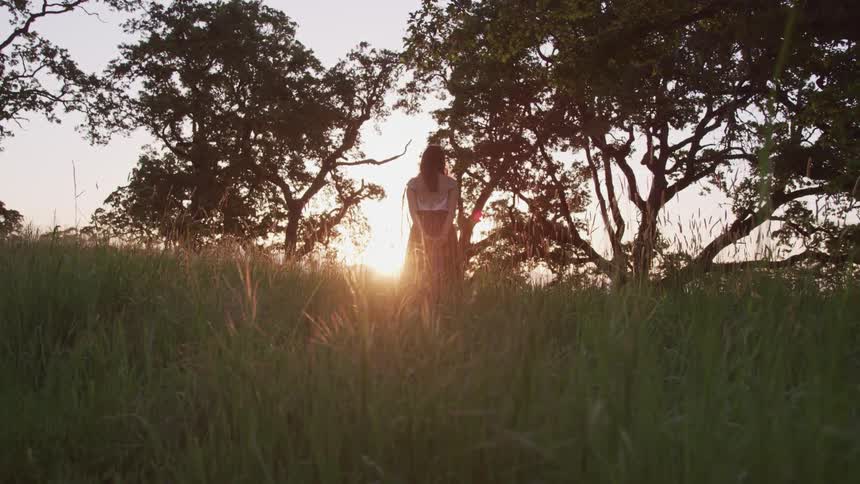A woman stands atop a grassy hill as the sun is low in the sky