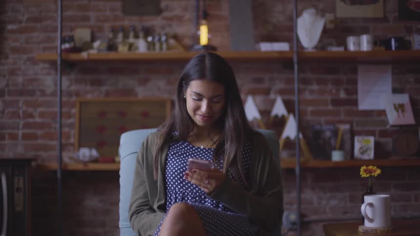 A woman sits in a cafe with a coffee mug looking at her phone and smiling