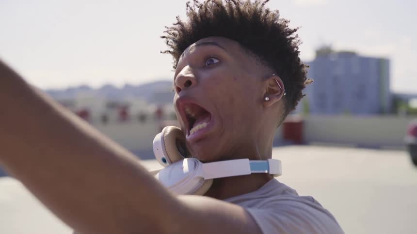 A young man uses his smartphone to take selfies while he makes funny faces as he stands at the top of a tall parking garage in a metropolis on a sunny day