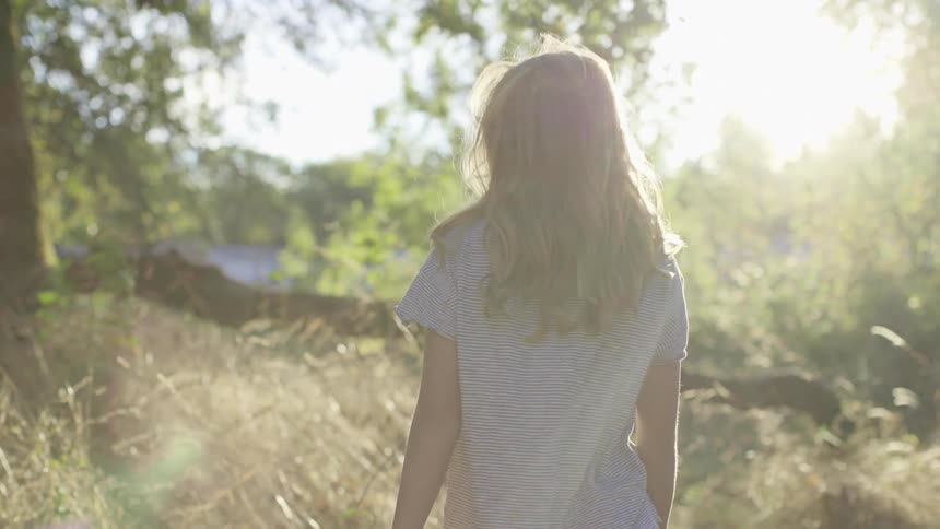 Girl with long hair walks through tall weeds into woods on a bright sunny day