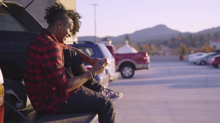 A young man sitting on the back of a vehicle in a parking lot in the evening smiles while using his smartphone