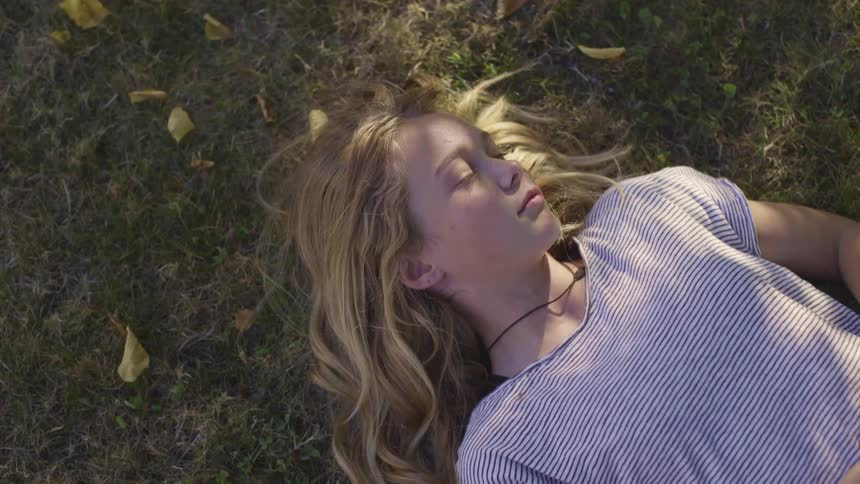 Young girl laying down in grass and leaves scattered about on the ground closes her eyes and smiles