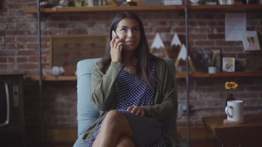 A woman sits on a chair in a room while talking on a phone