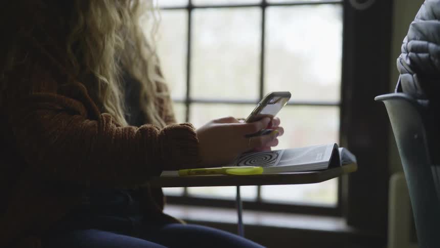 A teenage girl sits at her desk in school and texts on her cell phone