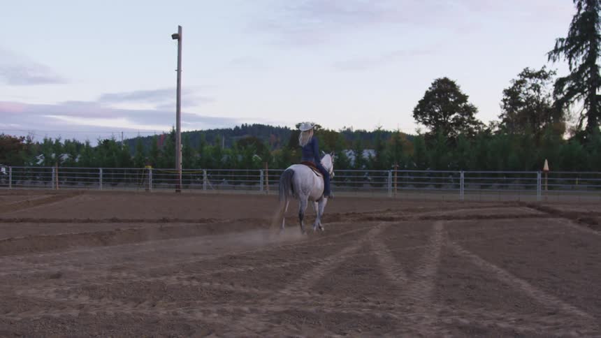 Blonde woman wearing cowboy hat rides horse inside a corral at evening