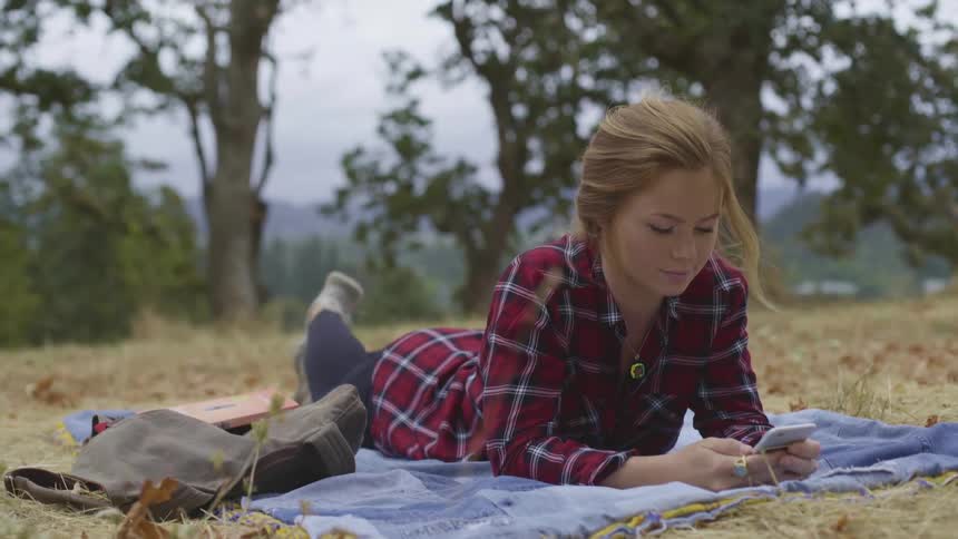 Young girl lays on her stomach outside on a blue blanket while texting on her phone at daytime