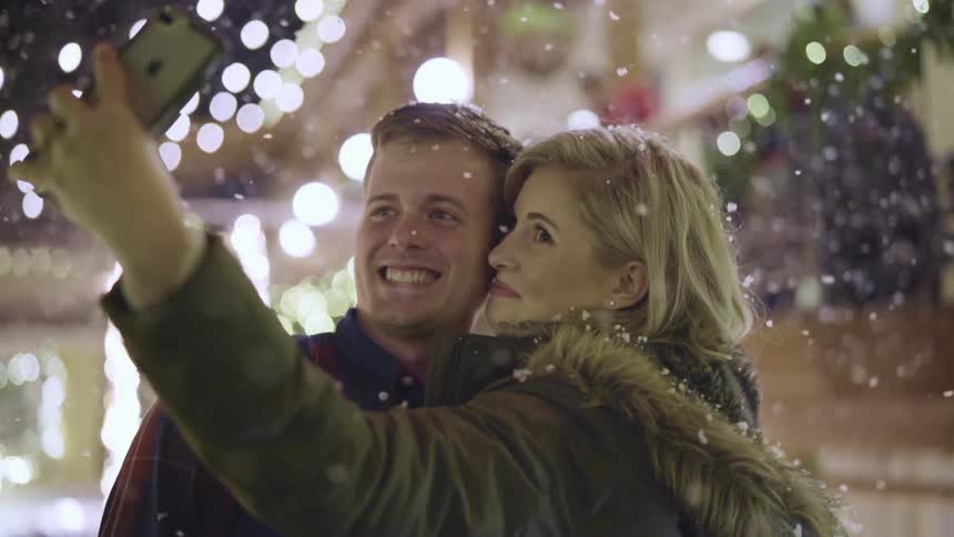 Woman snaps a photo of her and her boyfriend outdoors at an event with snow falling at night
