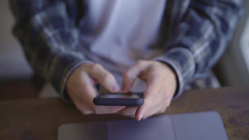 A man sitting down at a wooden table holds his smart phone while typing a message on it