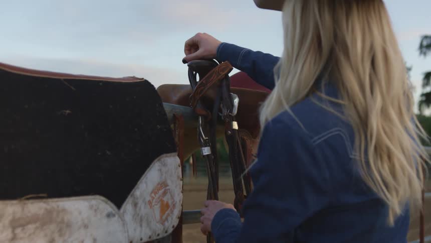 A woman sets the reins on top of a saddle resting on a fence