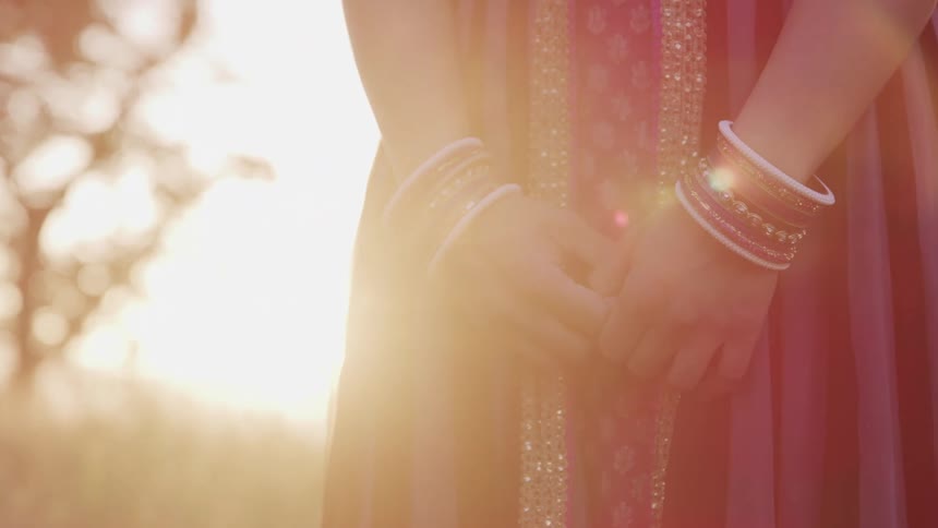 A woman in an indian style dress stands in a field with her hands clasped together