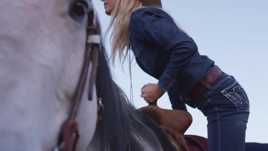 A young woman climbs into the saddle of a white horse under a clear blue sky
