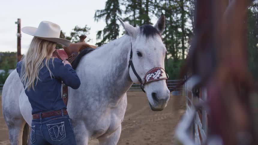 Woman adjusts a brown saddle on top of a white horse in a ranch enclosure outdoors at daytime