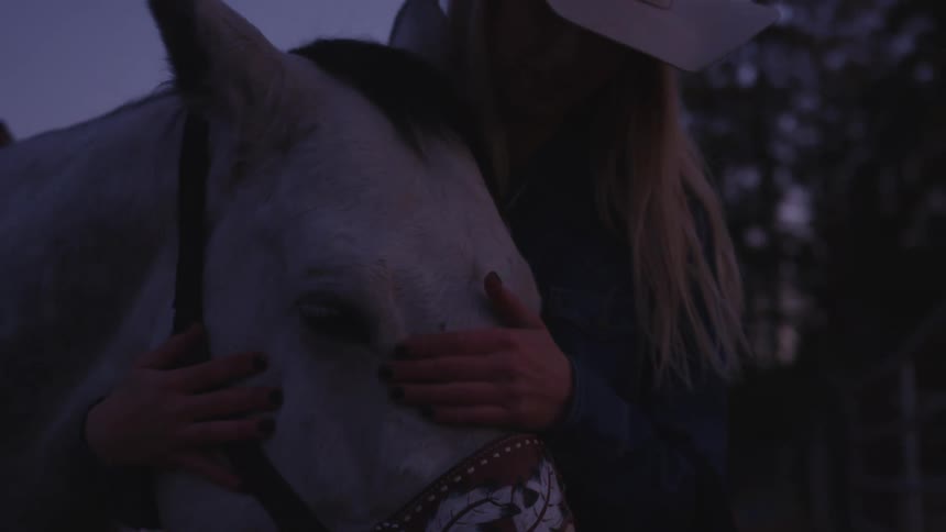 A blonde woman pets a horse while standing outdoors at night in front of trees