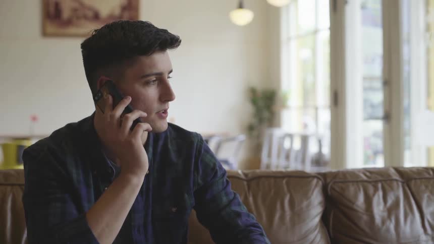 A male teenager sits on a leather brown couch talking on a cell phone indoors during the day
