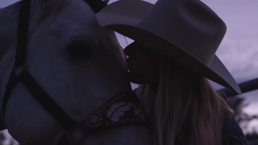 Woman wearing cowboy hat kisses a horse in the forehead at evening
