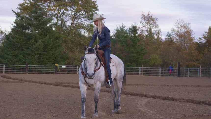A woman in a cowboy hat and head to toe denim climbs aboard a white board in a fenced arena