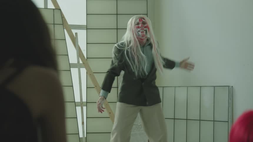 Man with red and white face paint and a white wig performing dance movements in an art studio while people watch