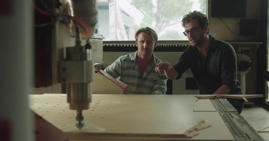 Two man supervising a machine carving a piece of plywood in a workshop