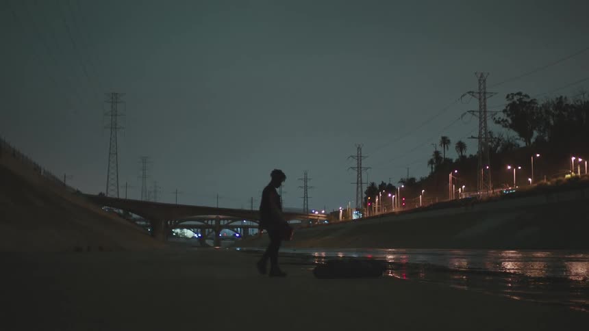Man walks over to a pile in a river trench in a city at night and pours liquid from a bucket on top