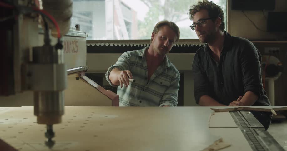 Two men sitting in front of machine with a wooden slab in doors having a discussion at daytime