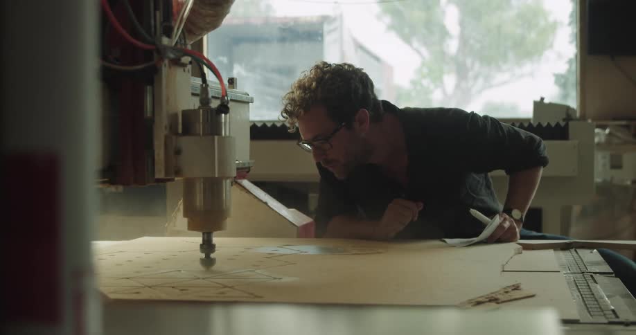 A machine on a table carves a pattern into a slab of thin wood and a man sitting next to it blows off the dust