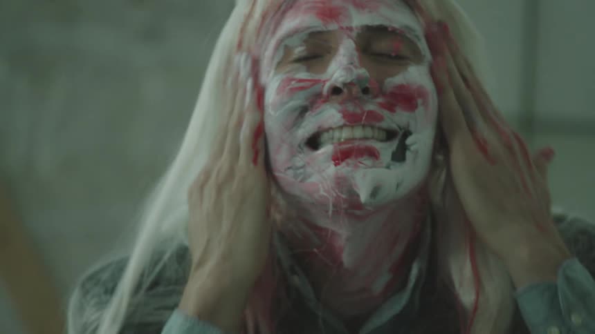 A man in a blonde wig sits on the floor in a room undergoing construction and wipes face paint on his face with a wide smile