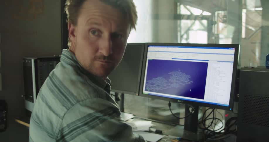 A man using a computer in an office turns to look behind him for a moment before returning his gaze to his screen