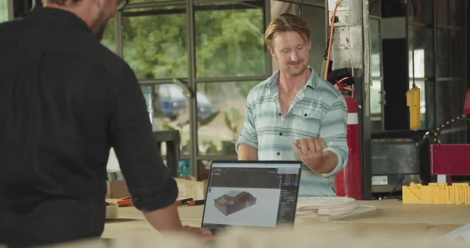 Two men working in a wood workshop while one looks at a design of a home on a laptop computer