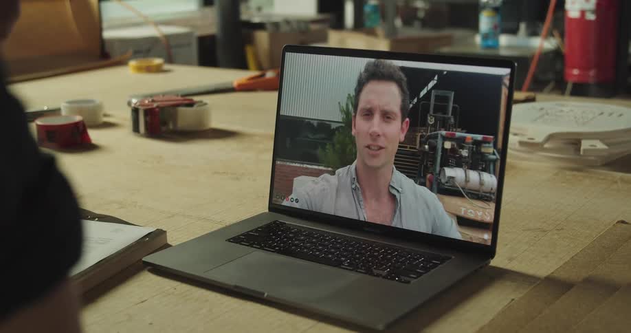 A person in a workshop uses a laptop to wave at a man through video chat