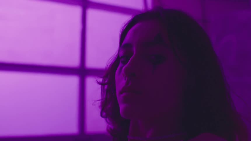 A woman is lying on her back in a bare room with a window in purple light