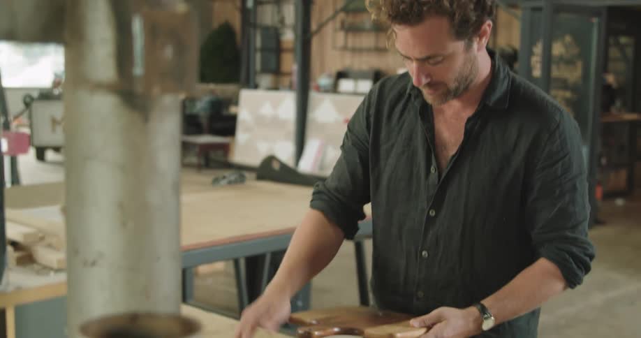 A man stands at a table in a workshop brushing off a wooden paddle