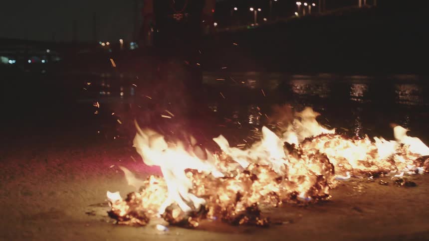 Young man stands in front of a smoldering fire in a city channel at night