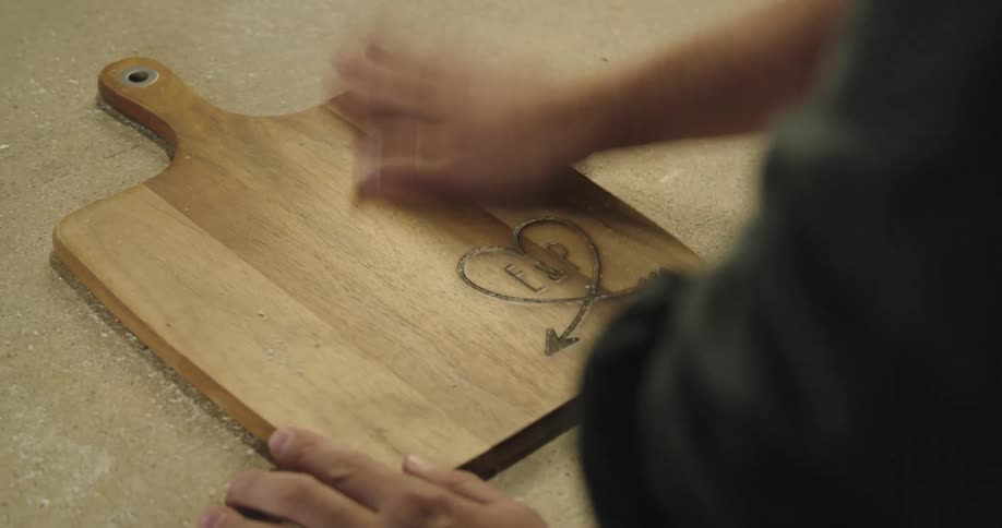 A man wipes saw dust off of a cutting board he engraved