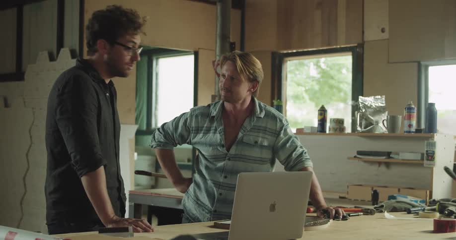 Two men stand in a room under construction and talk to someone on their laptop