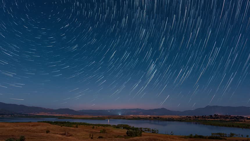 Landscape with mountains and a river surrounded by fields and a night sky with stars turning into streaks