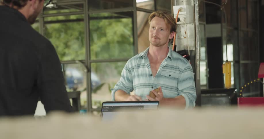 A man polishes a piece of woof in his hand while he talks to a man across from him in a workshop at daytime