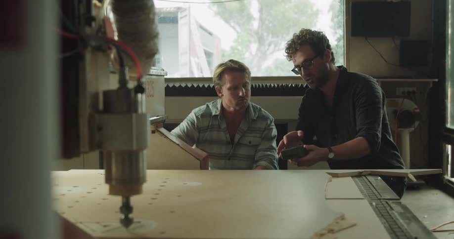 One man watches holes being drilled in plywood in a factory as another man shows him a tool